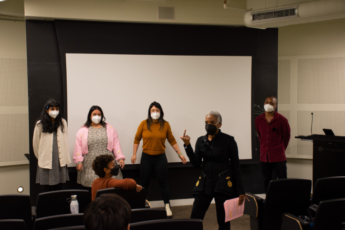 Karen Proctor teaching to a group in front of a white screen with students behind her participating in her exercise. Ryana Burrell is sitting in front of her, and David Proctor is standing behind her.
